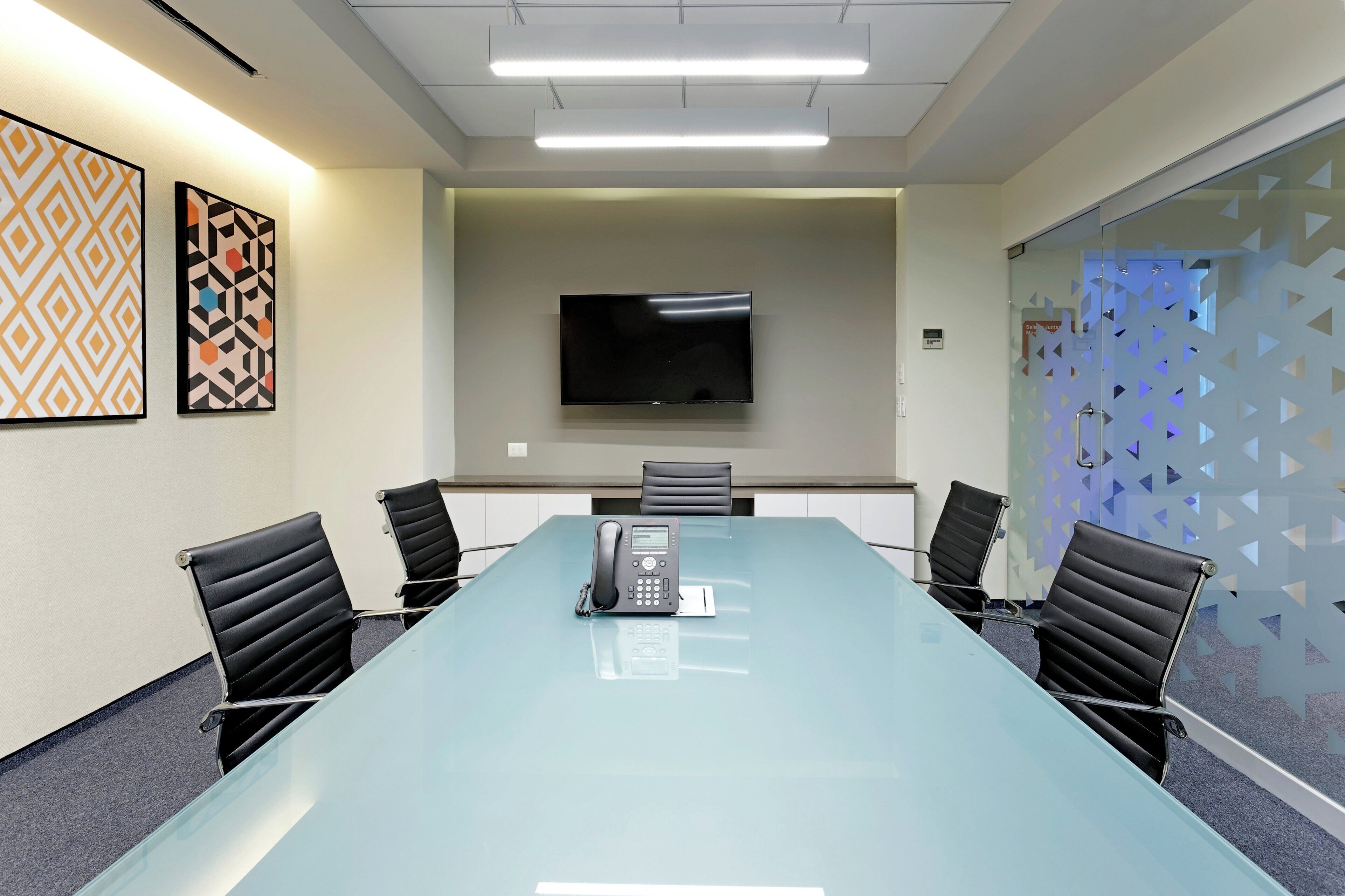 Un salón de conferencias con una mesa de vidrio, cuatro sillas, un televisor de pantalla plana montado en la pared y dos cuadros enmarcados.