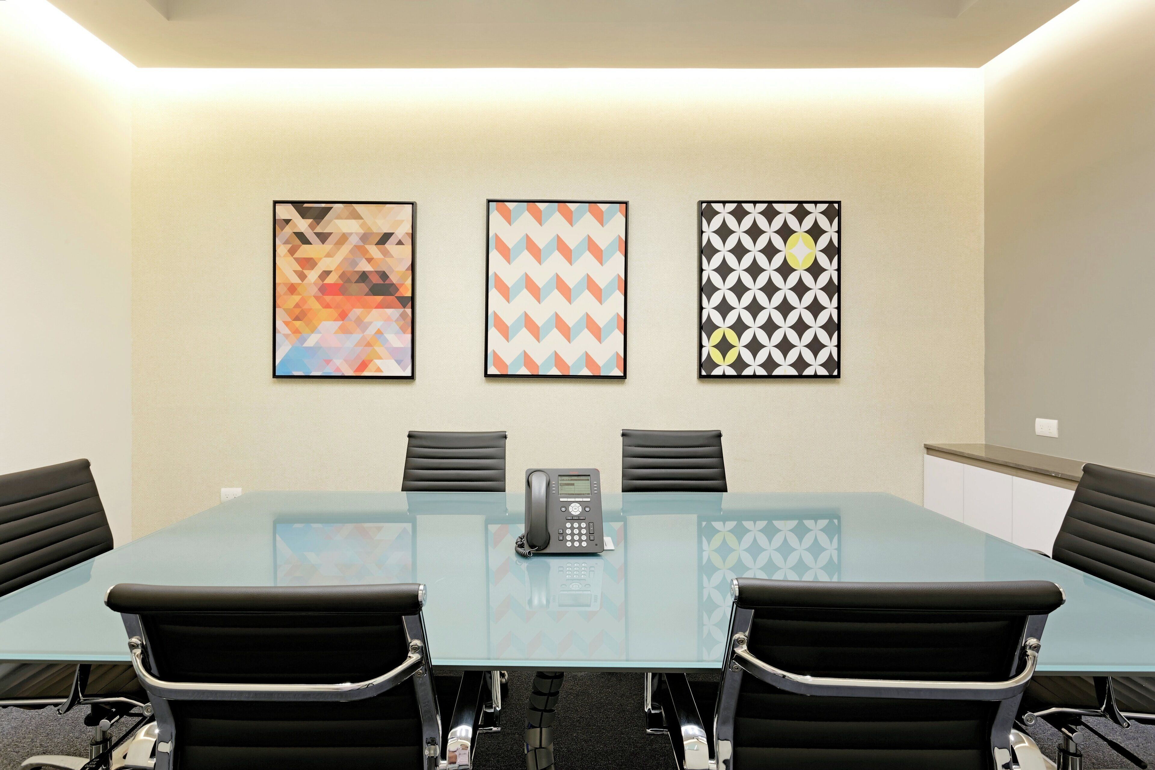 A conference room with a glass table, four black chairs, a telephone, and three framed artworks on the wall.