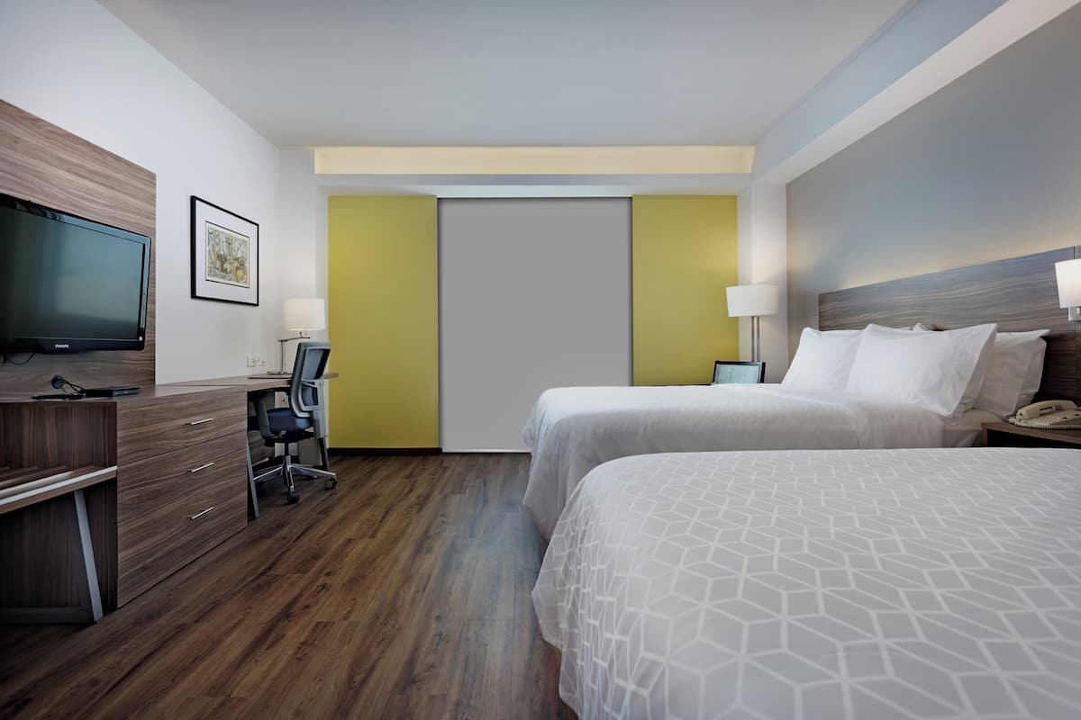 Une chambre d’hôtel avec un bureau en bois, une télévision, deux lits et une porte surmontée d’un mur d’accent jaune.