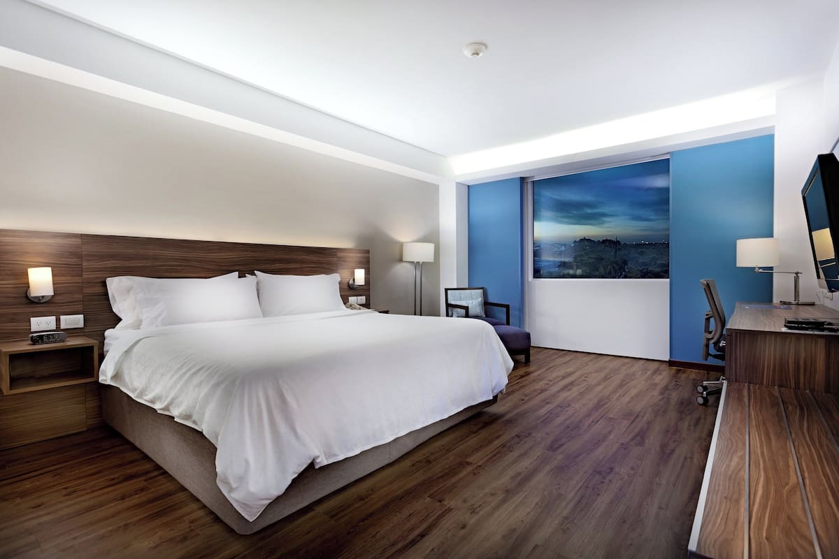 Une chambre d’hôtel moderne avec un grand lit, un plancher en bois, une table de chevet avec une lampe, un bureau avec une chaise et une grande fenêtre donnant sur le paysage urbain.