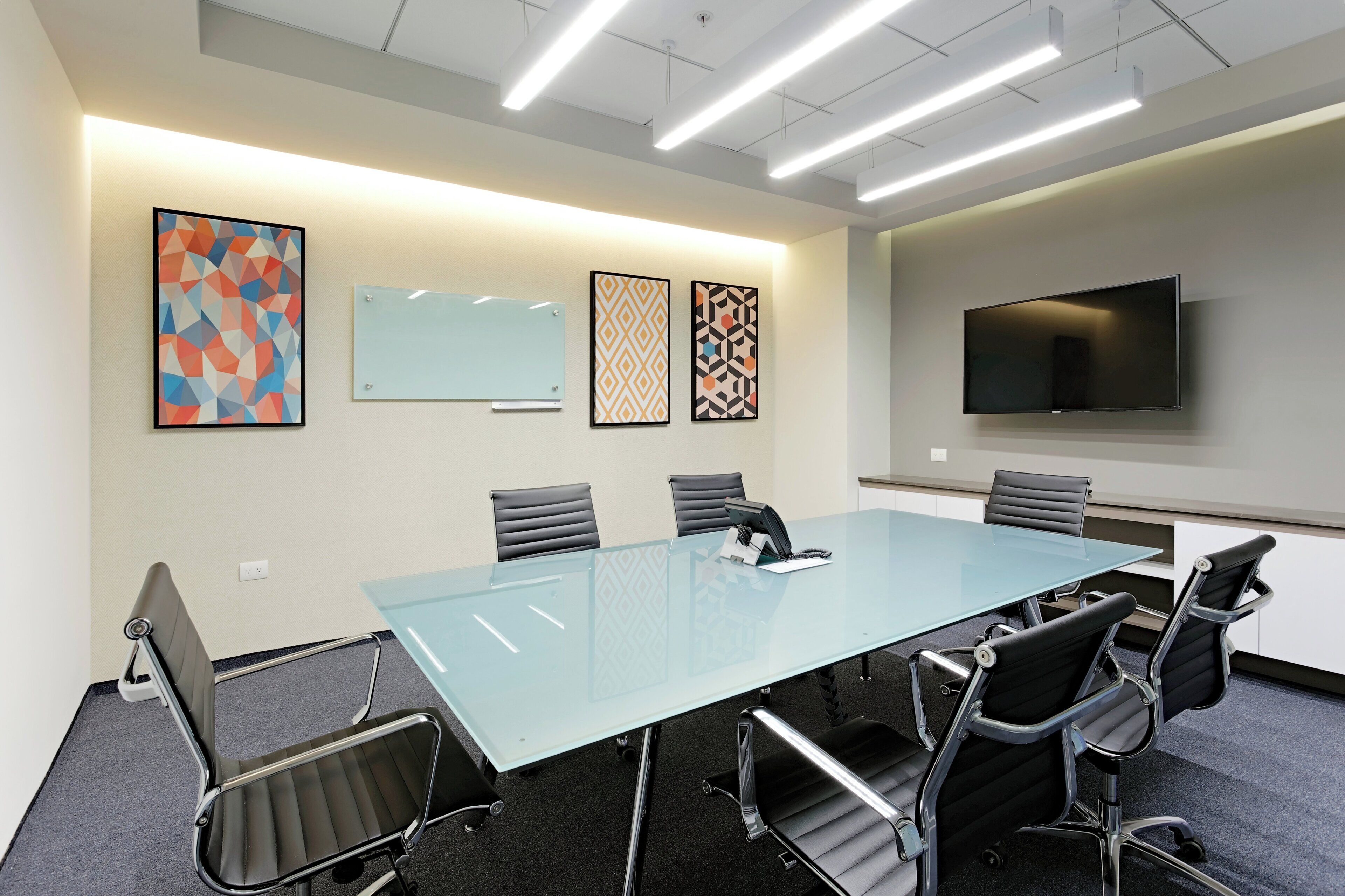 A conference room with a glass table, six chairs, a whiteboard, and a flat-screen TV on the wall.