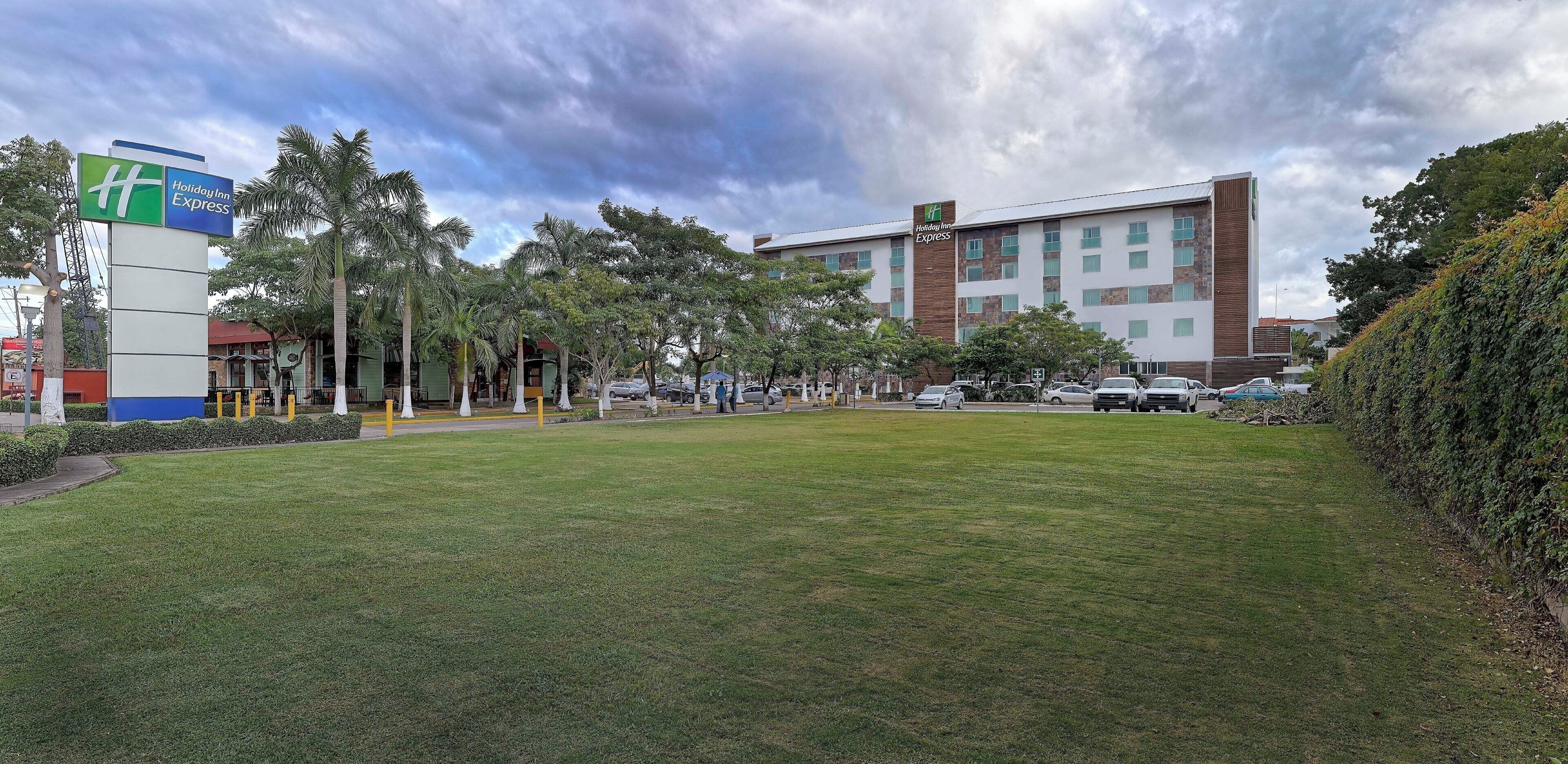 Un hotel Holiday Inn Express con un patio cubierto de césped, palmeras y un estacionamiento.
