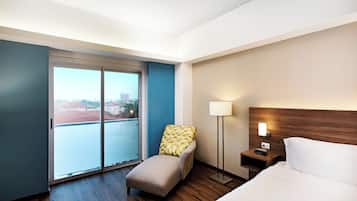 Habitación de hotel con cama, mesita de noche, lámpara, silla y un amplio ventanal corredero que da a un balcón con vista a la ciudad.
