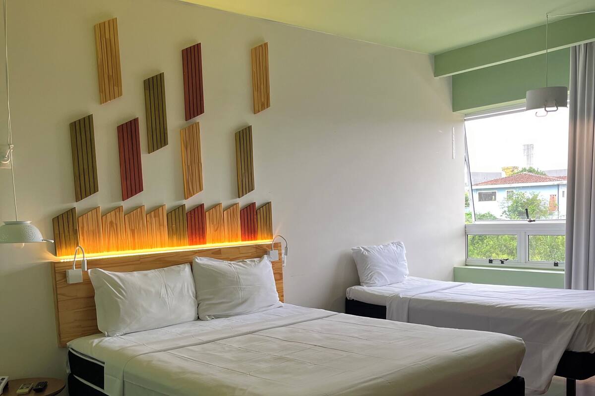 Hotelkamer met twee bedden, een houten hoofdbord, een nachtkastje met een lamp en een raam met uitzicht op gebouwen.