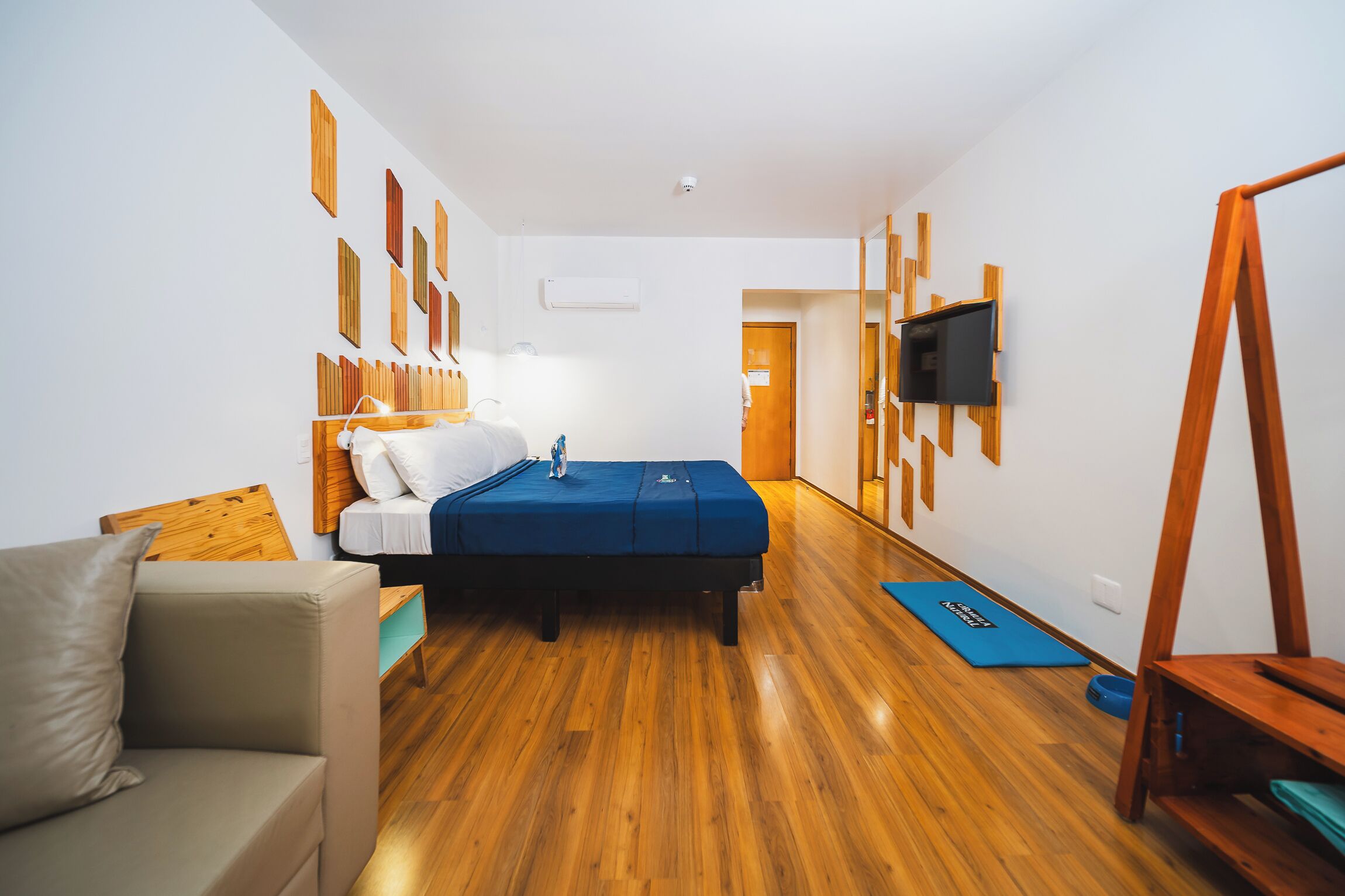 A compact hotel room with a bed, a sofa, a TV mounted on the wall, and a wooden floor.