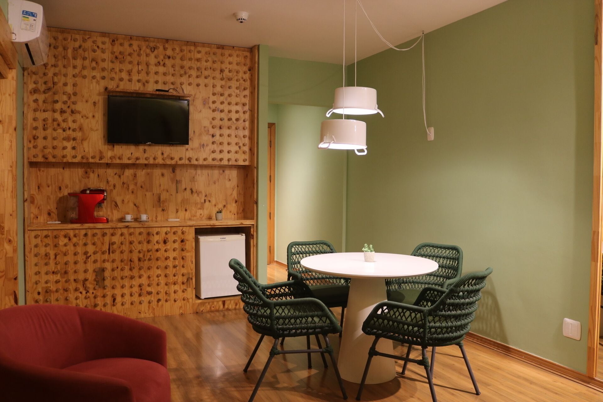 A room with a wooden wall, a flat-screen TV, a small refrigerator, a red coffee machine, a white table with four green chairs, and a hanging light fixture.