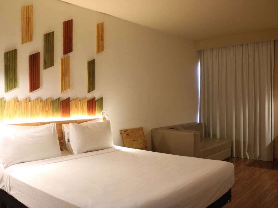 Quarto de hotel com uma cama grande, cabeceira de madeira, sofá e janela com cortinas.