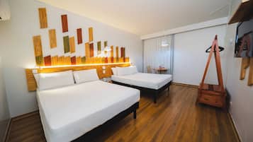 A hotel room with two beds, a wooden floor, a wall-mounted light fixture, and a small table with a chair.
