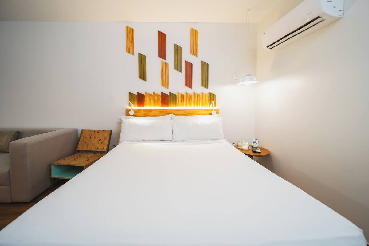 Een hotelkamer met een groot bed, een houten hoofdeinde, een wandairconditioner, een nachtkastje met een lamp en een klein houten bijzettafeltje.