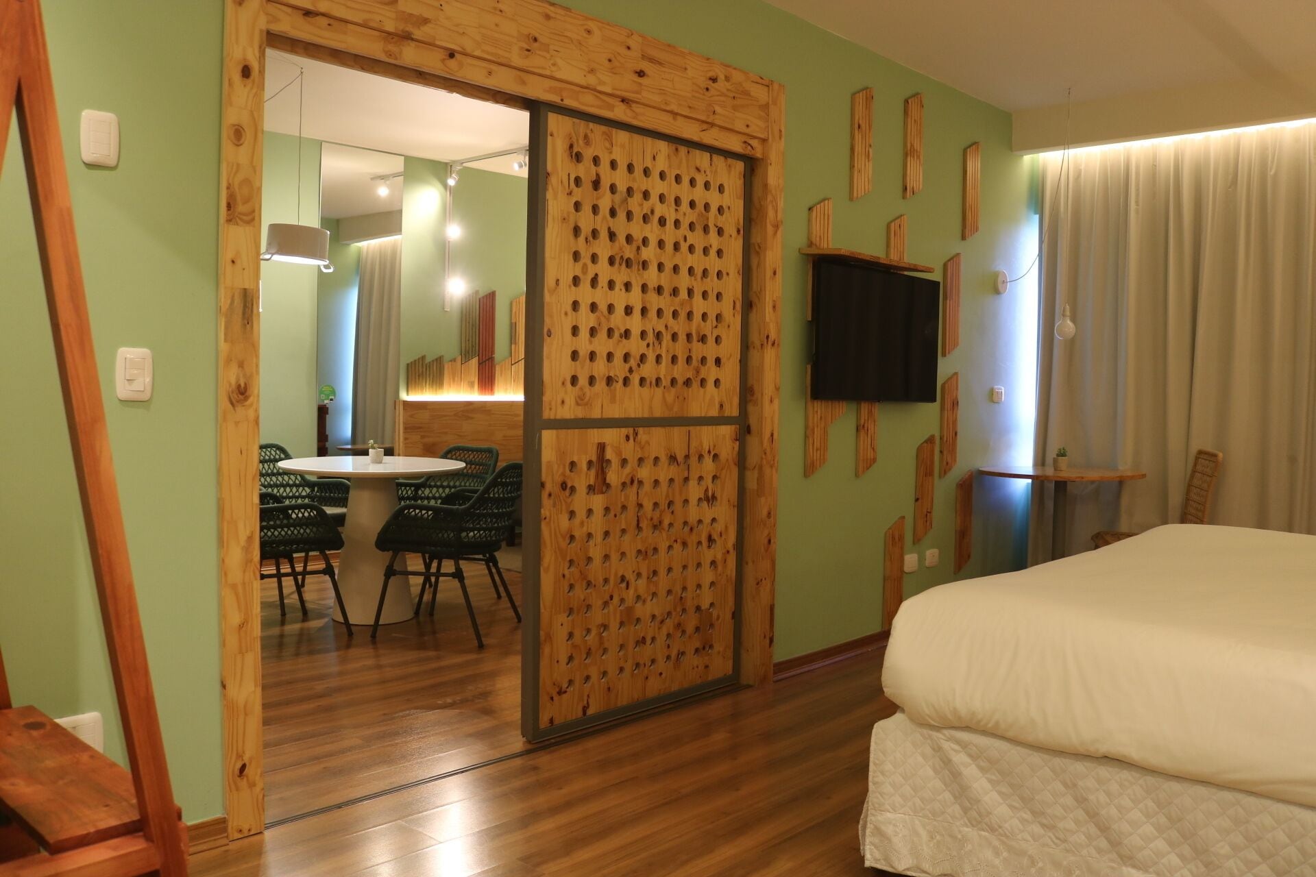 Una habitación con cama, un separador de madera con agujeros, una zona de comedor con sillas, un televisor en la pared y una ventana con cortinas.
