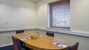 A conference room with a wooden table, four chairs, a projector screen, a window with blinds, and a power outlet on the wall. - Holiday Inn Express Leicester - City by IHG (Leicester)
