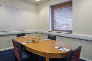 Une salle de conférence avec une table en bois, quatre chaises, un écran de projection, une fenêtre avec des stores et une prise de courant au mur.