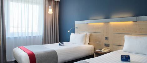 A hotel room with two beds, a bedside table with a lamp, a phone, and a window with curtains.
