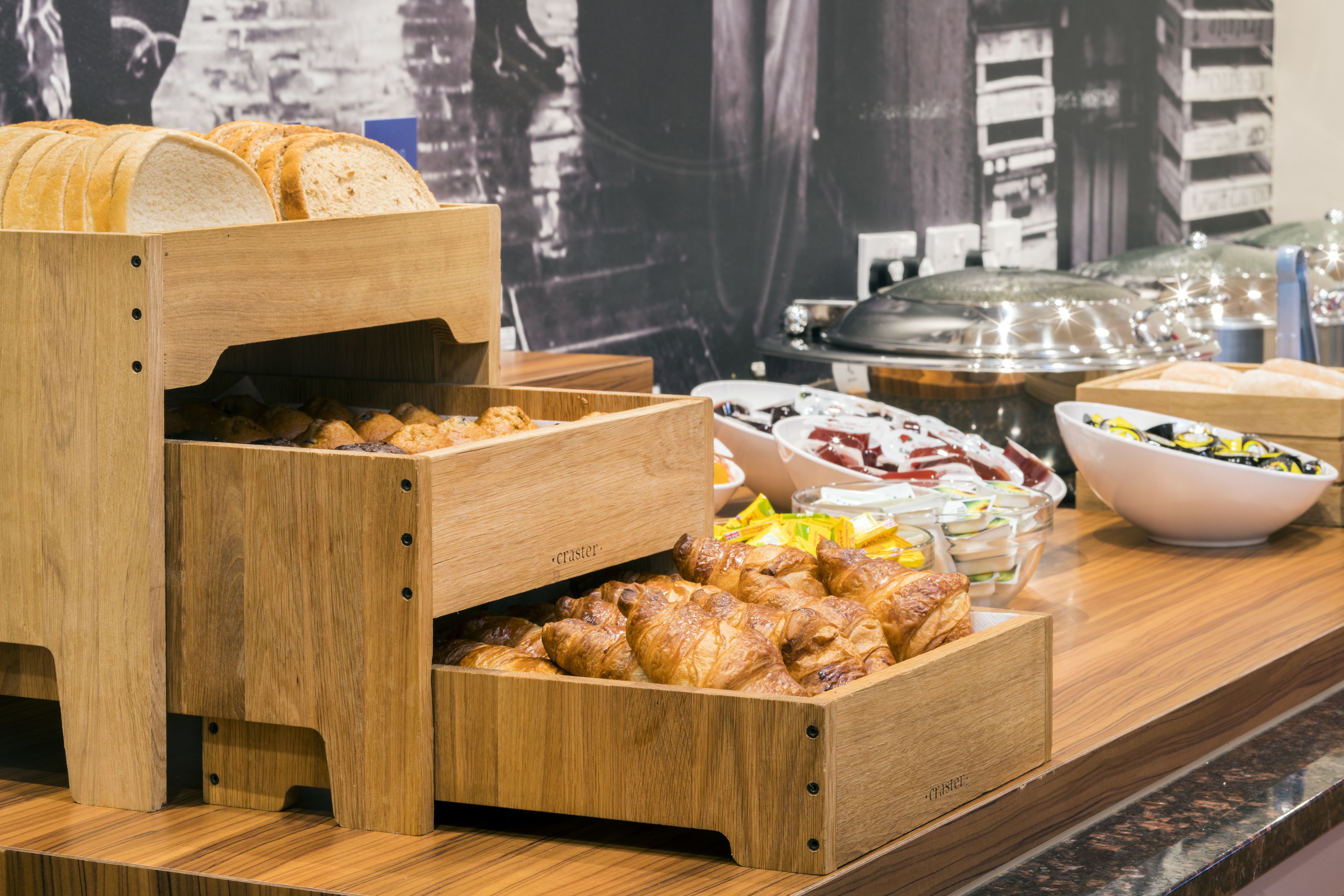 Una barra de buffet con diversas opciones de comida, incluyendo pan, pastelería y un tazón grande de ensalada.