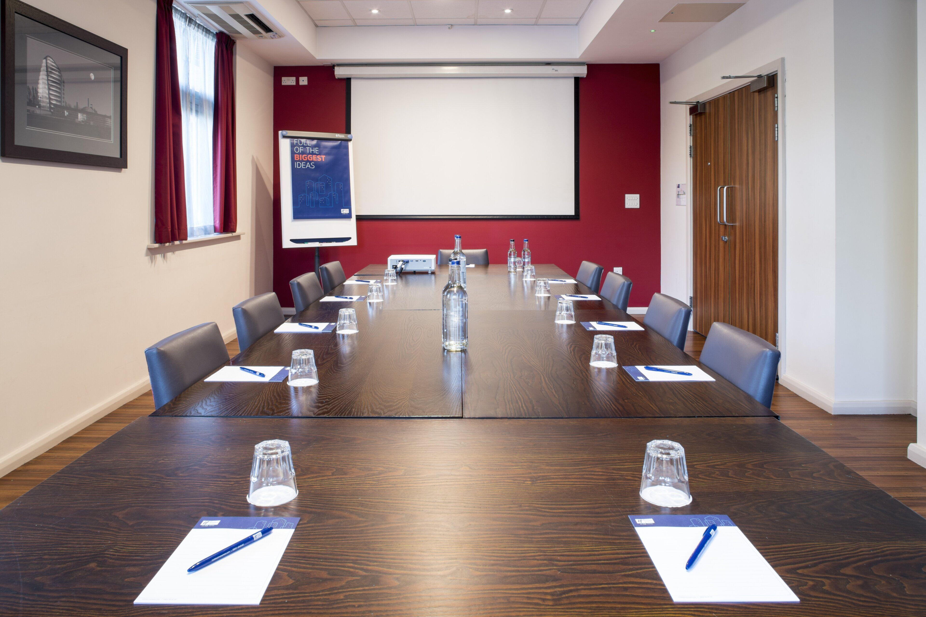 A conference room with a long table, chairs, water bottles, pens, and a projector screen.