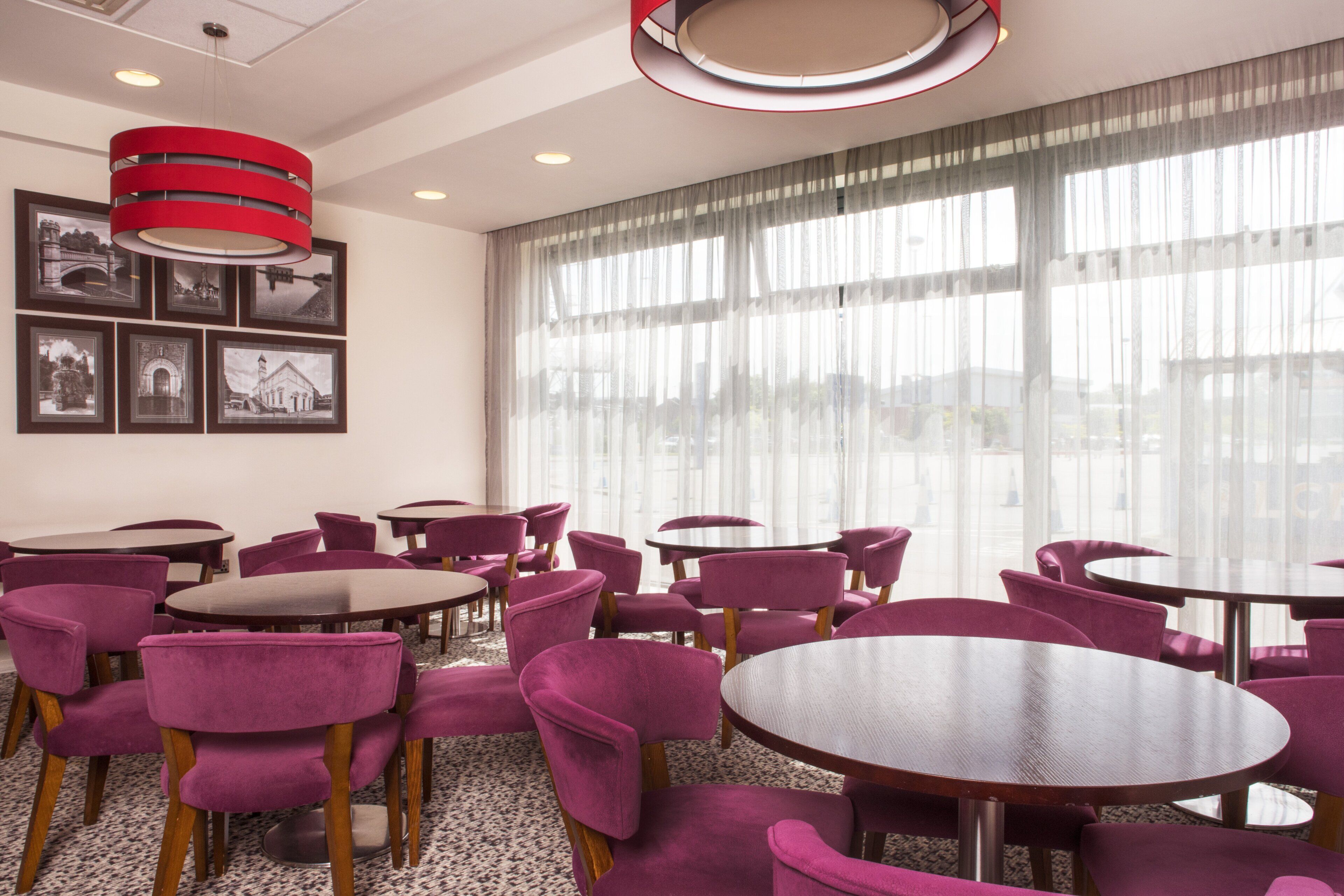 A hotel dining area with round tables and purple chairs, large windows with sheer curtains, and a red pendant light.