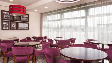 A hotel dining area with round tables and purple chairs, large windows with sheer curtains, and a red pendant light.
