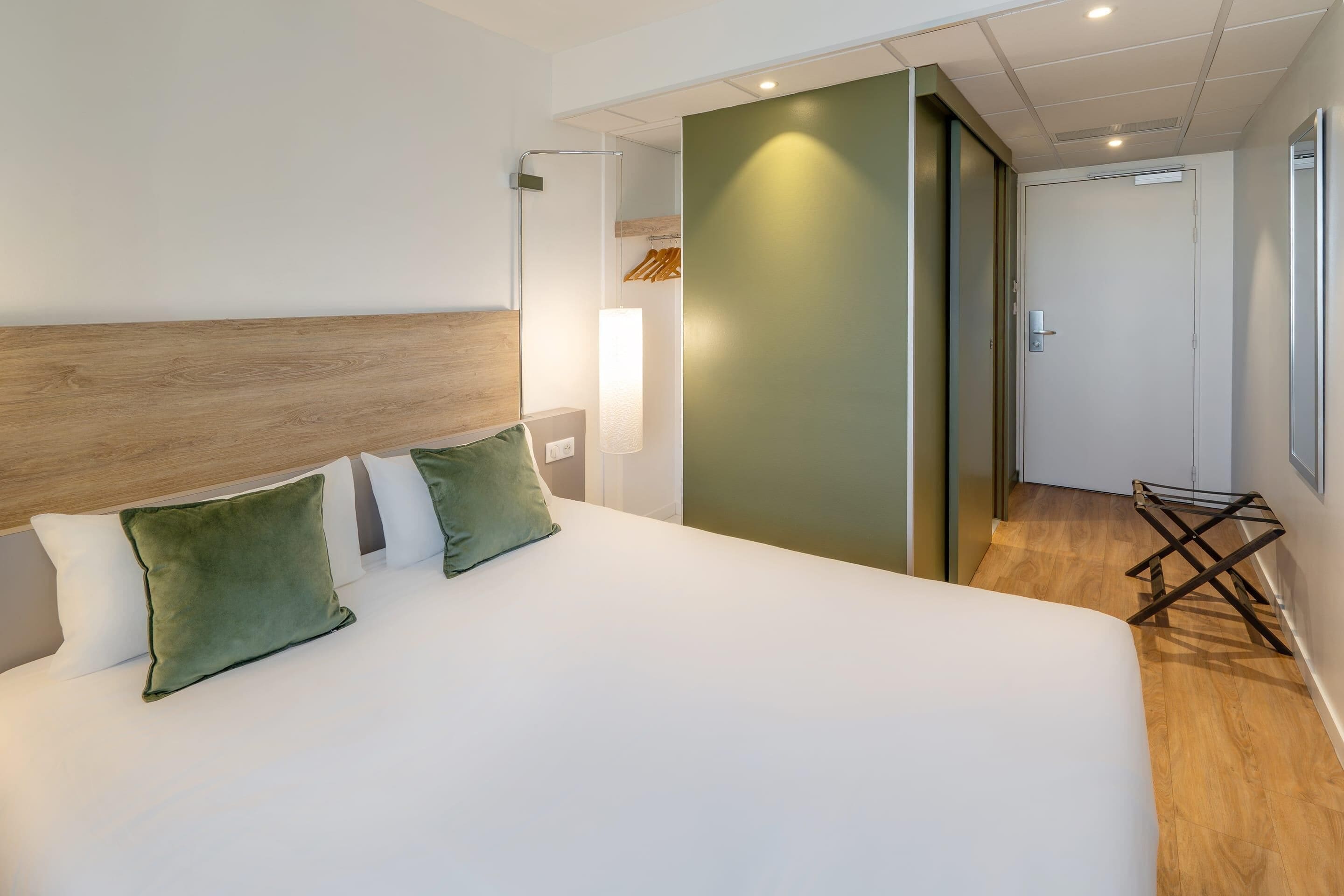 Habitación de hotel con una cama grande, cabecera de madera, pared verde decorativa, un pequeño rincón de descanso y una puerta que da a otra habitación.