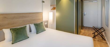 Habitación de hotel con una cama grande, cabecera de madera, pared verde decorativa, un pequeño rincón de descanso y una puerta que da a otra habitación.