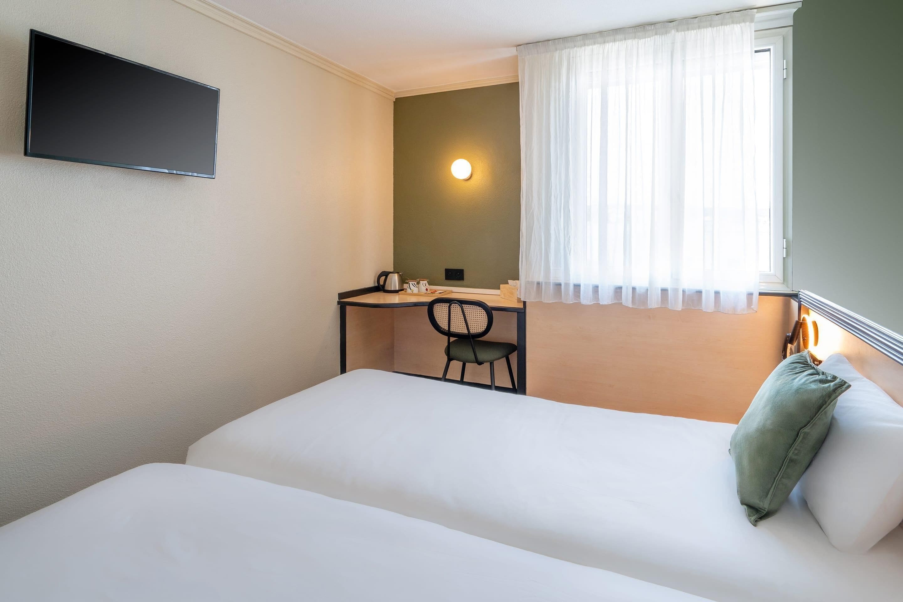 Een hotelkamer met een bed, een bureau met stoel, een televisie, een raam met gordijnen en een wandlamp.