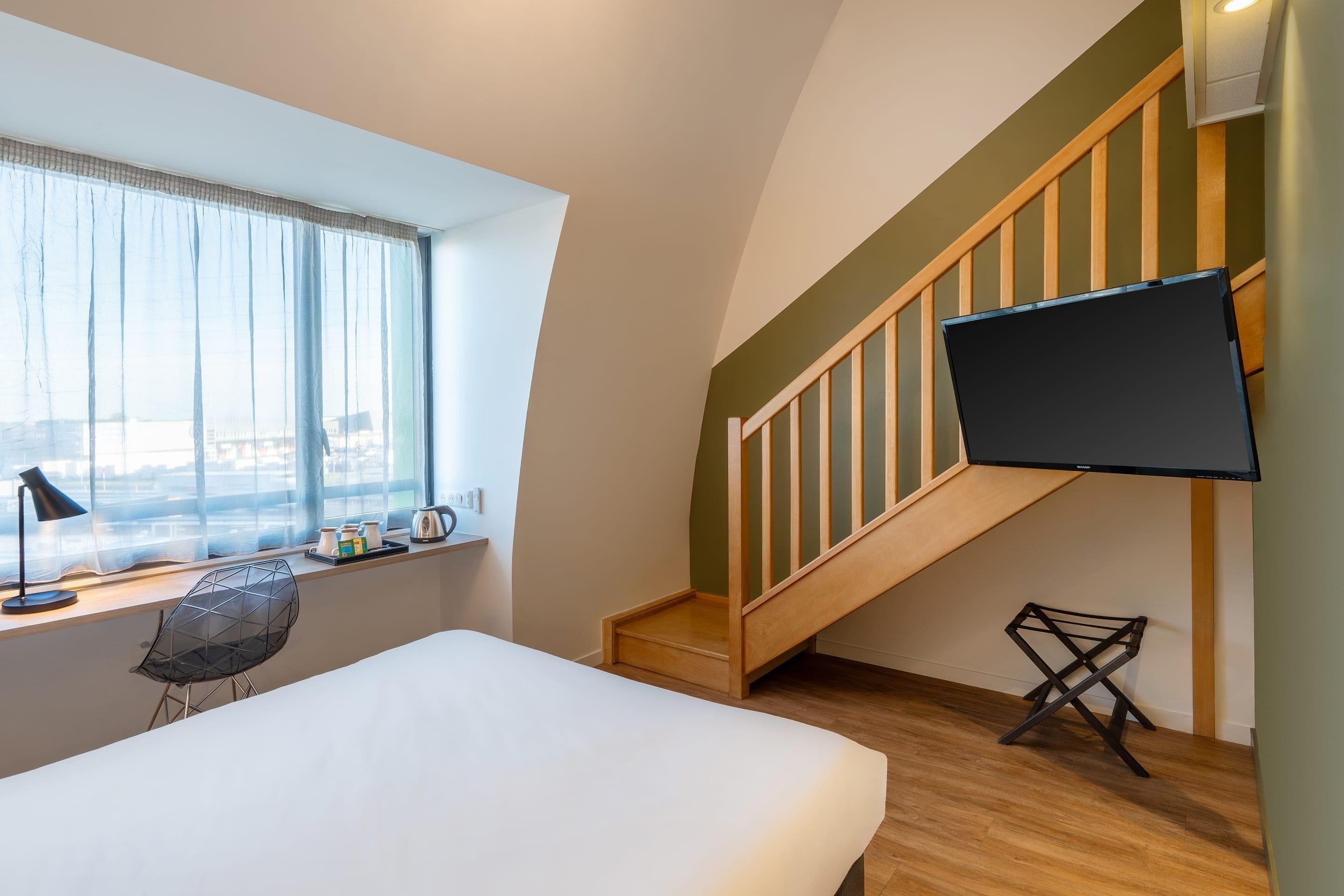 A bedroom with a bed, a desk with a chair, a TV mounted on the wall, and a staircase.
