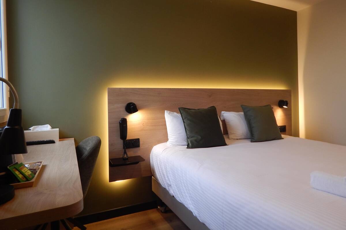 Une chambre d’hôtel avec un lit bien fait, un bureau avec une chaise, et une table de chevet avec une lampe et une boîte de mouchoirs.