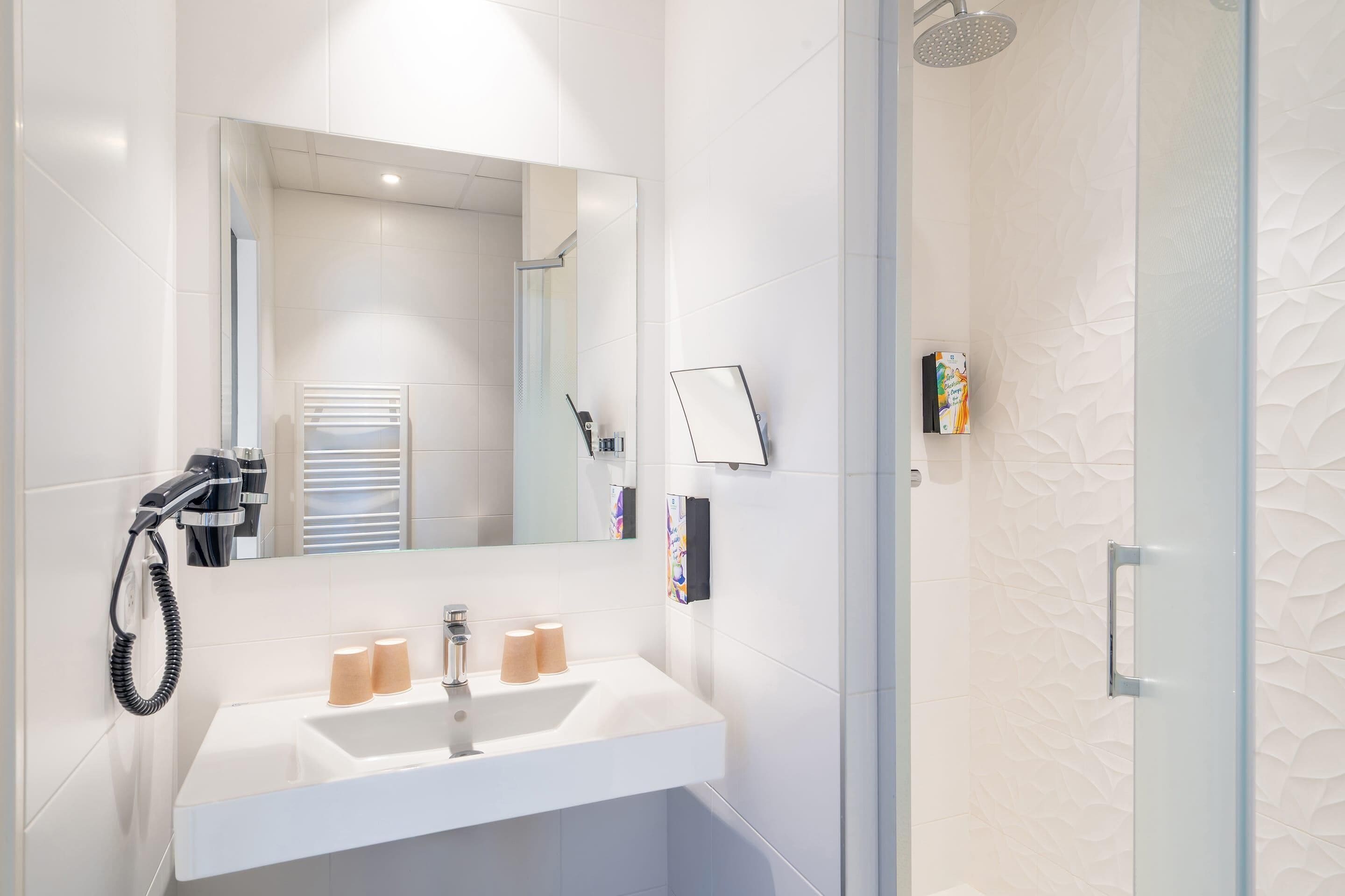 Une salle de bain moderne avec un lavabo blanc, un miroir, un sèche-cheveux et une cabine de douche.