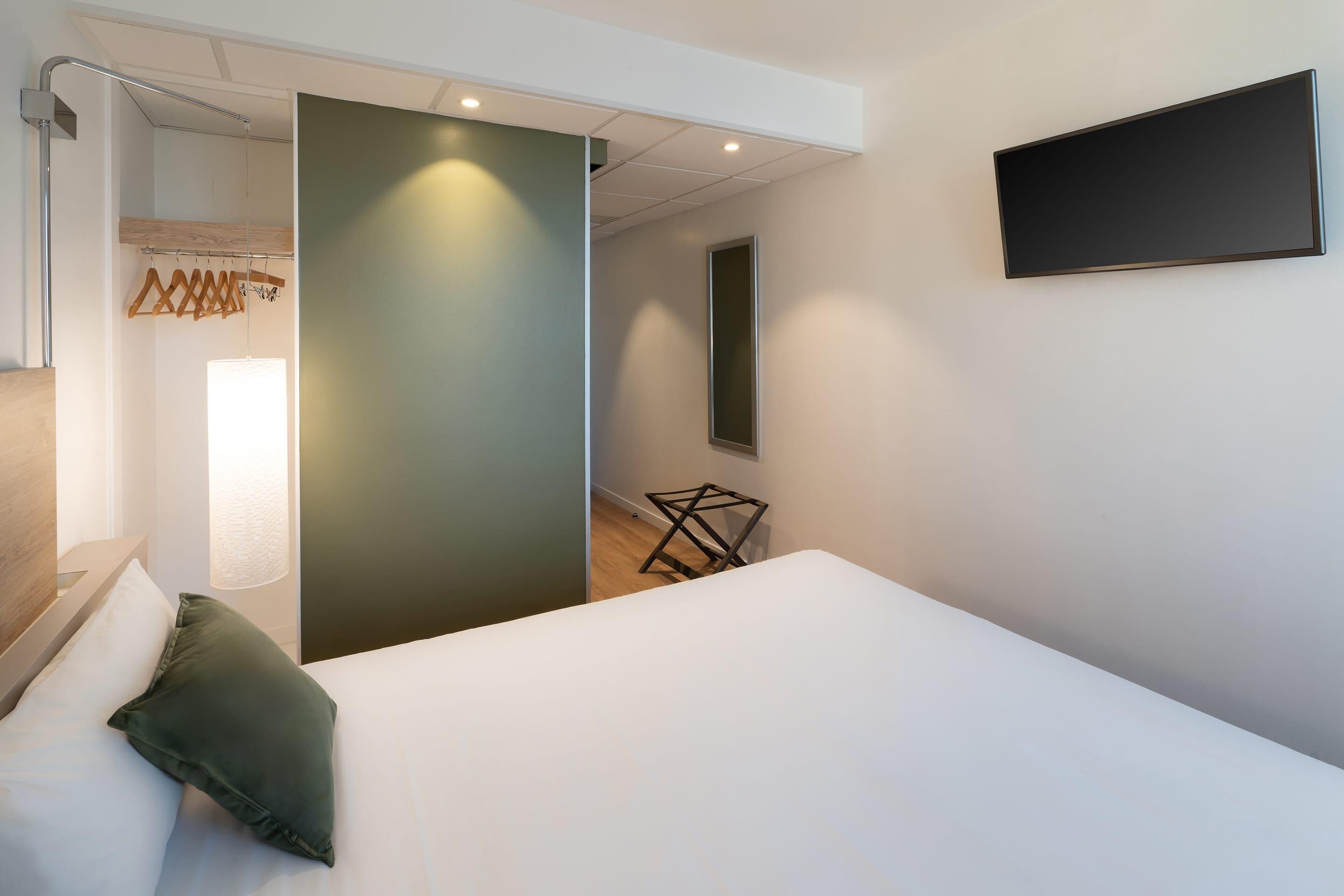 A modern hotel room with a bed, a hanging coat rack, a wall-mounted TV, and a small table.