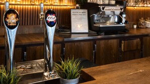 Een bar met twee biertaps, een hangende lamp en een toog met een koffieapparaat en menukaart.