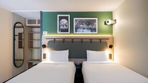 A hotel room with two beds, a mirror, a shelf unit, and two framed pictures on the wall.