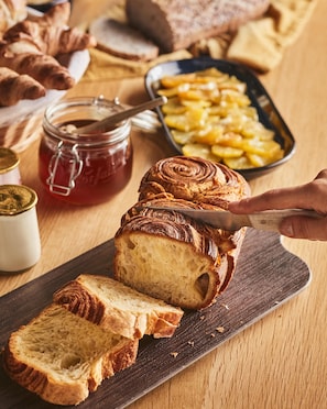 A loaf of bread with a swirl pattern on a wooden cutting board, accompanied by a jar of honey and a bowl of sliced potatoes.