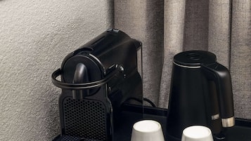 Un mini-bar dans une chambre d’hôtel, avec une machine à café, deux tasses blanches et divers en-cas et boissons.