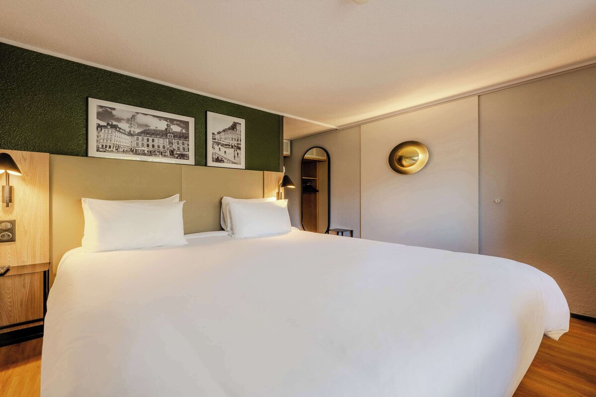 Une chambre d’hôtel avec un grand lit, deux lampes fixées au mur, une tête de lit en bois et deux photographies en noir et blanc accrochées au mur.