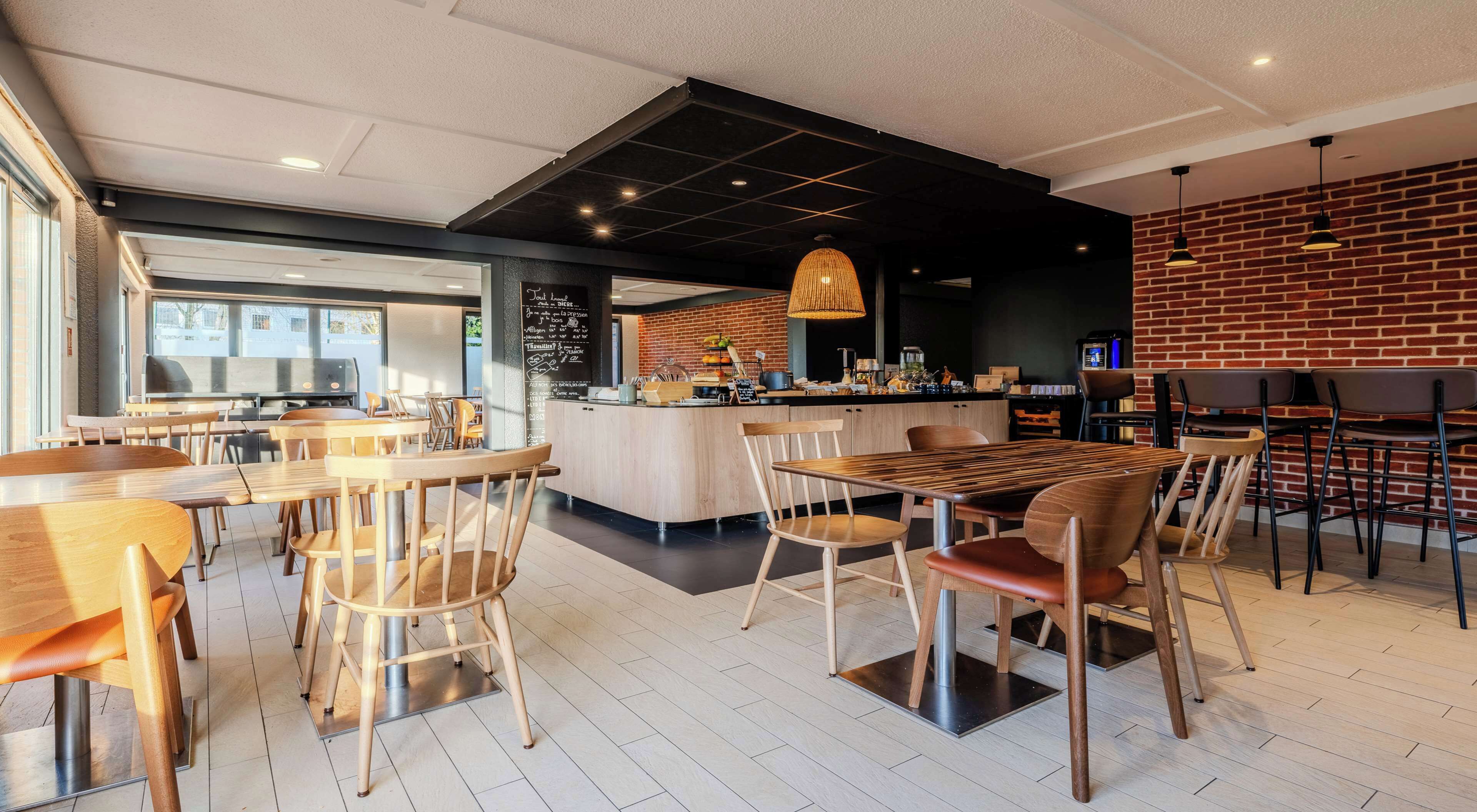 Ein modernes Restaurantambiente mit Holztischen und -stühlen, einer zentralen Bar und einer Wand mit Backsteinverkleidung.