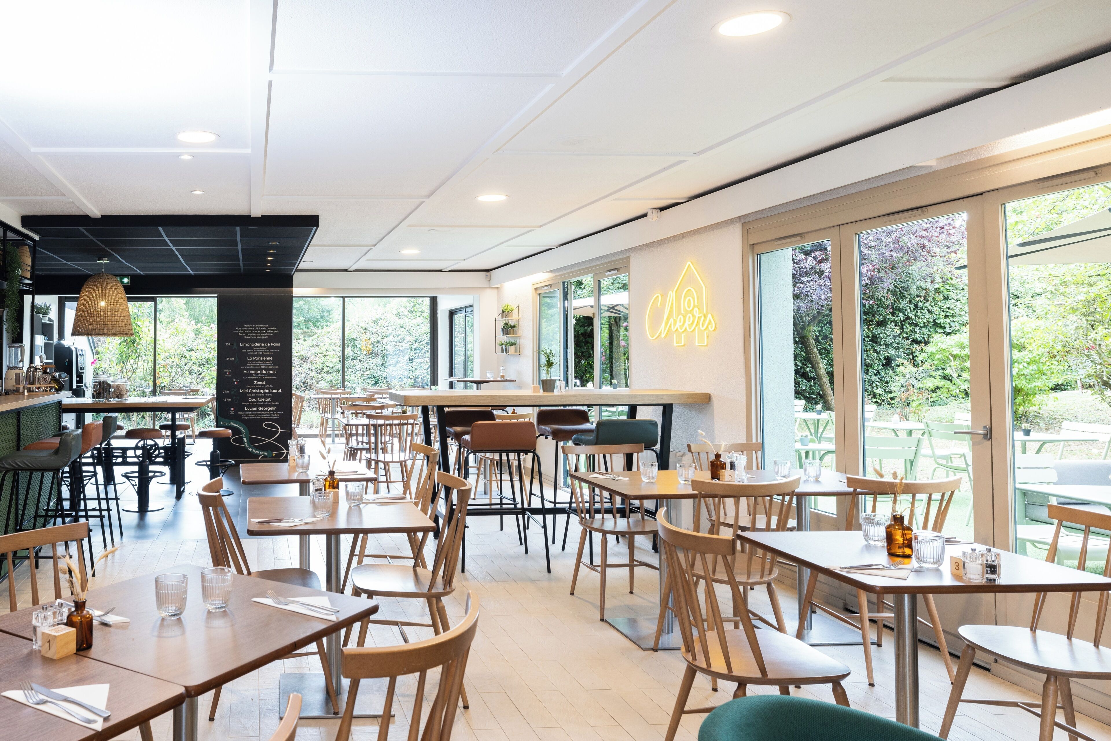 Un restaurante moderno con mesas y sillas de madera, un letrero de neón y amplias ventanas con vistas a zonas verdes.