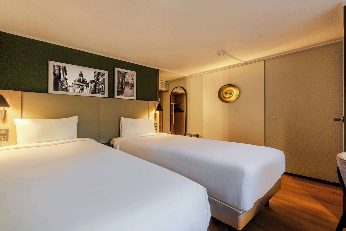 Une chambre d’hôtel avec deux lits, un miroir, un porte-manteau et deux tableaux encadrés au mur.