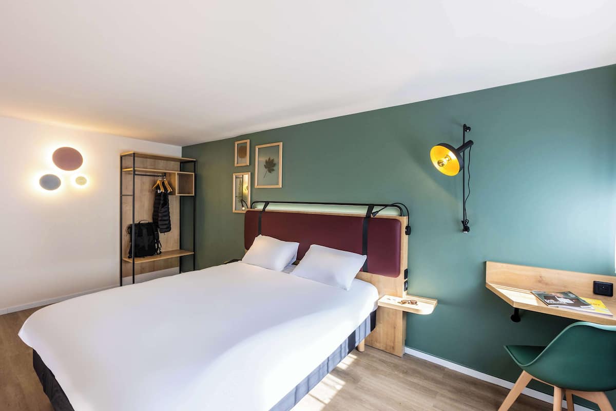 Une chambre moderne avec un grand lit, un bureau et une chaise. On y trouve une armoire avec des vêtements suspendus et une petite étagère.
