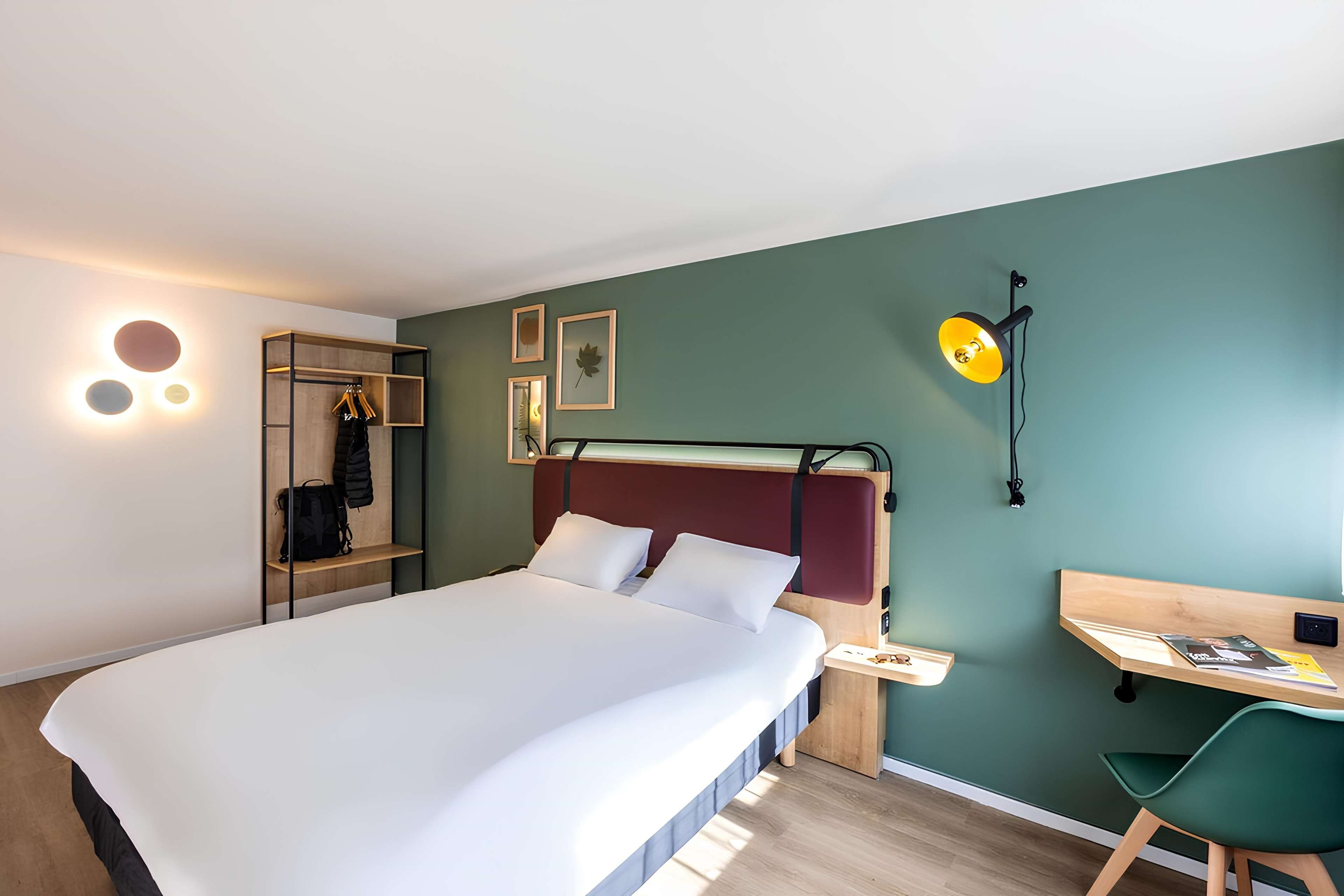 A modern hotel room with a bed, a desk, a chair, and a wardrobe.