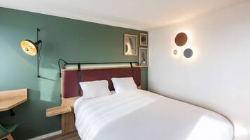 A neatly made bed with white linens, a desk with a chair, and a wall-mounted lamp.