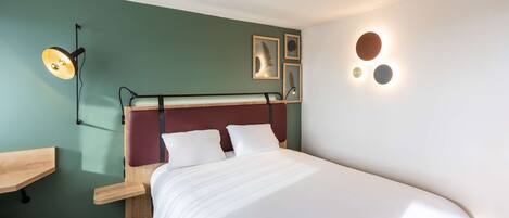 A neatly made bed with white linens, a desk with a chair, and a wall-mounted lamp.