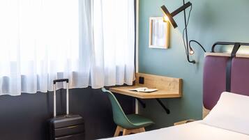 A hotel room with a bed, a desk, a chair, a suitcase, and a window with curtains.