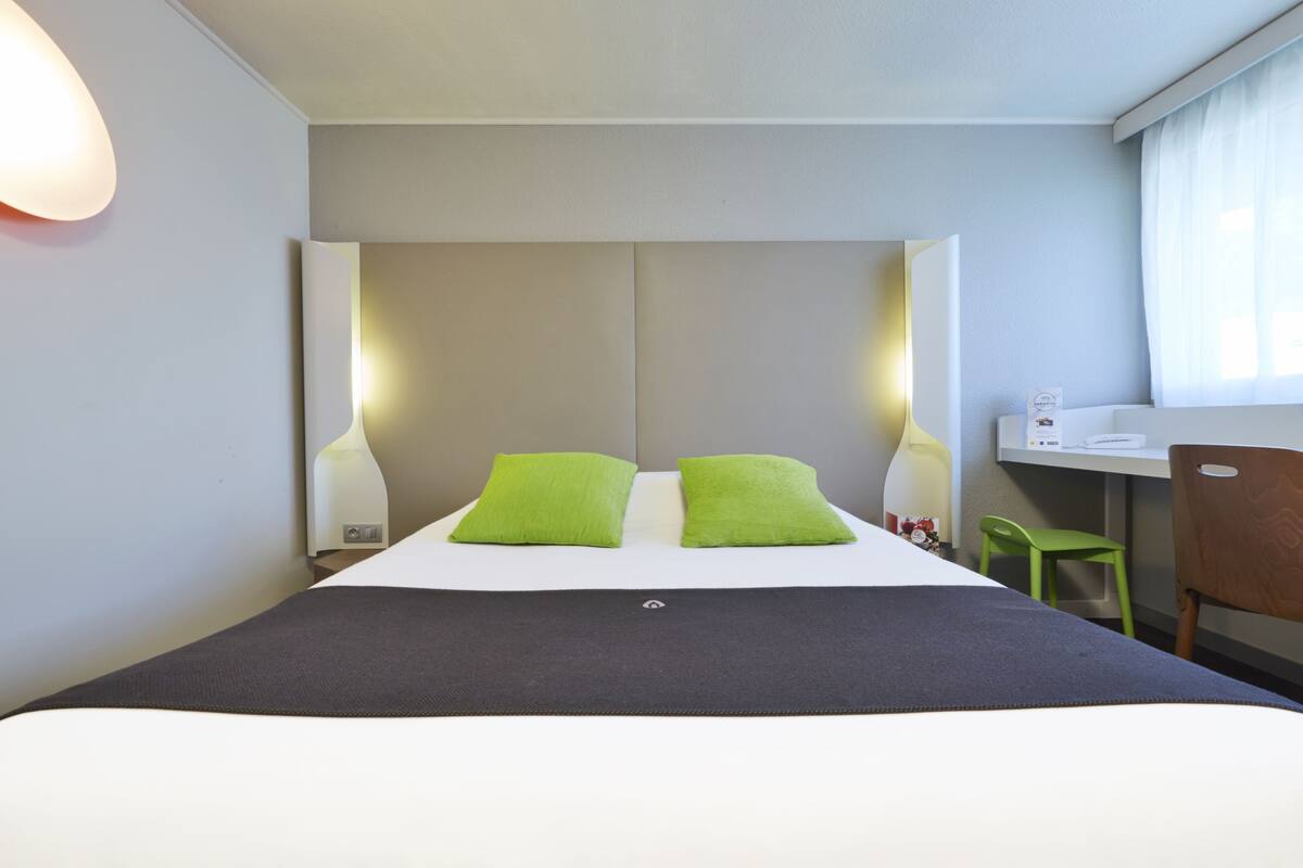 A hotel room with a bed, two green pillows, a desk, a chair, and a nightstand with a lamp.