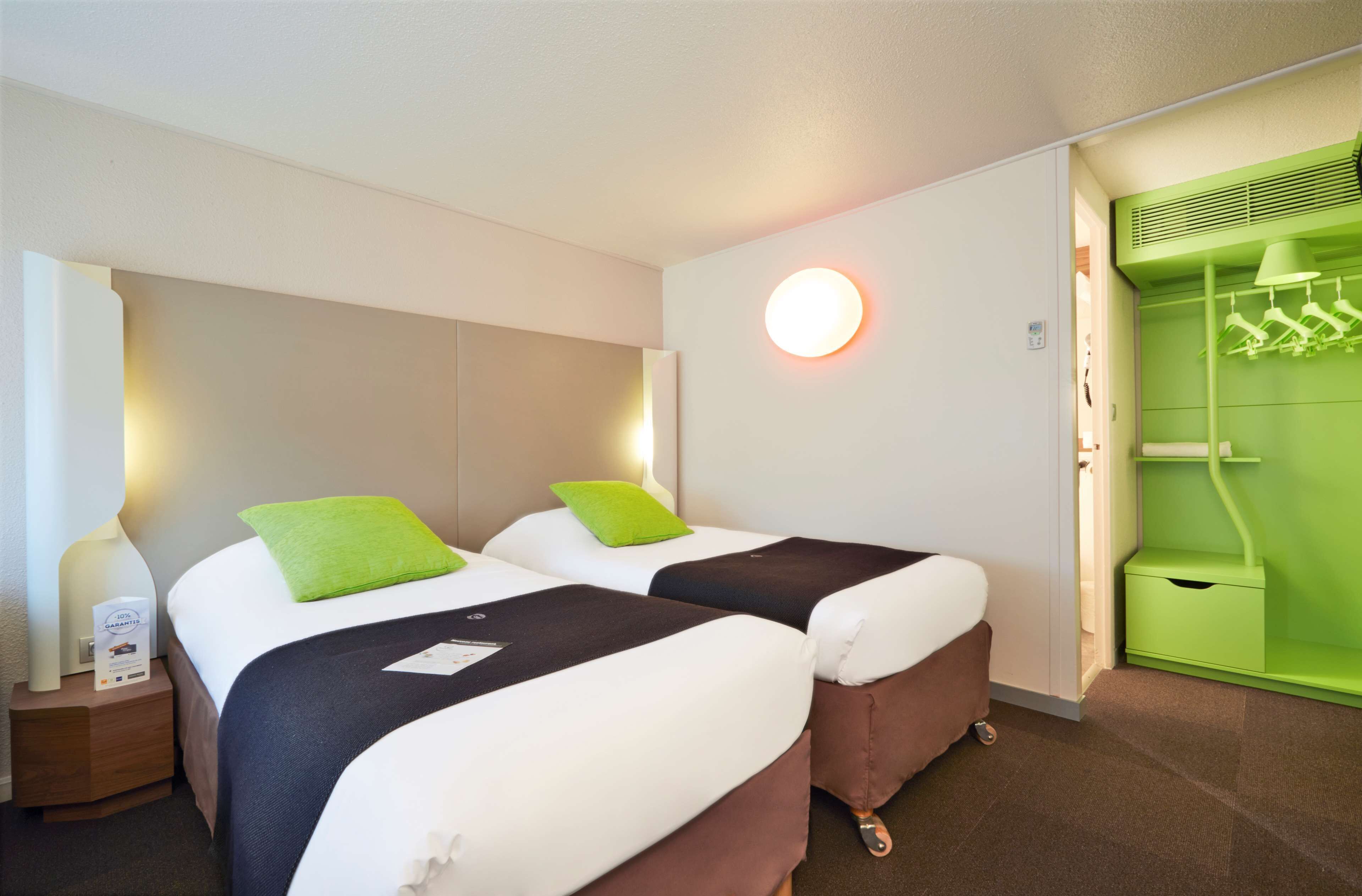 A hotel room with two beds, a green wardrobe, and a bedside table with a lamp.
