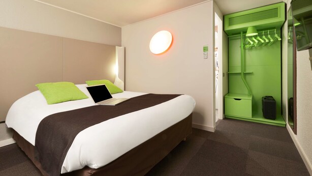 A modern hotel room with a neatly made bed, a green wall-mounted heater, and a green storage unit with hanging clothes. - Campanile L'Isle d'Abeau - Bourgoin Jallieu (L'Isle-d'Abeau)