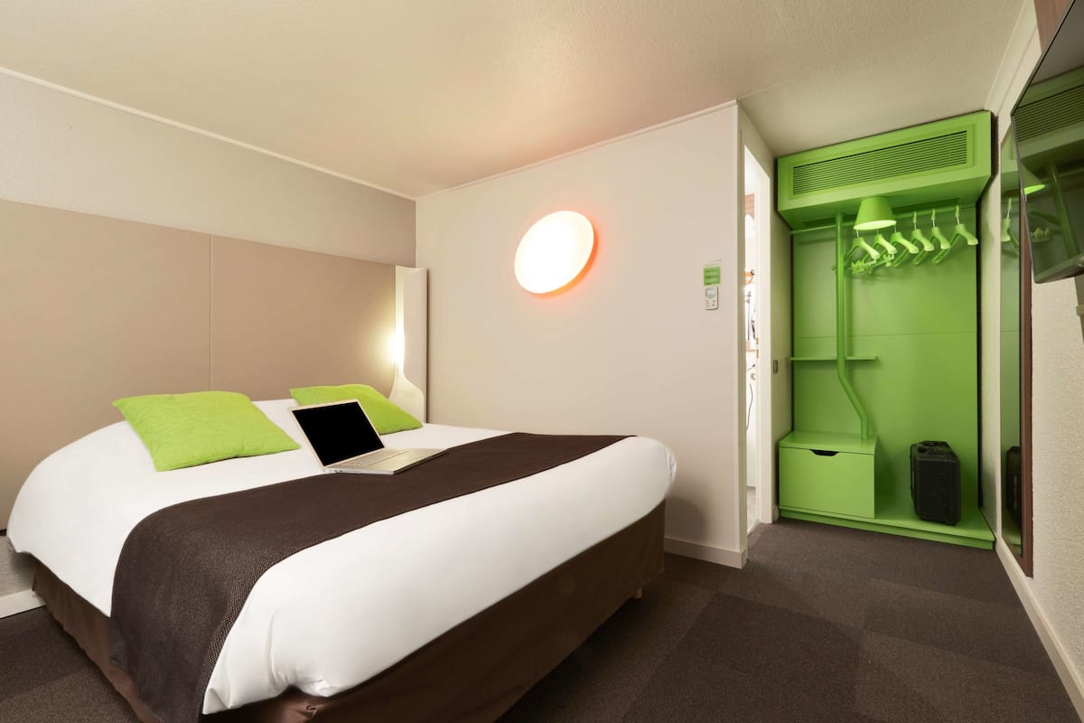 Une chambre d’hôtel moderne avec un lit soigneusement fait, un radiateur mural vert et un meuble de rangement vert où sont suspendus des vêtements.