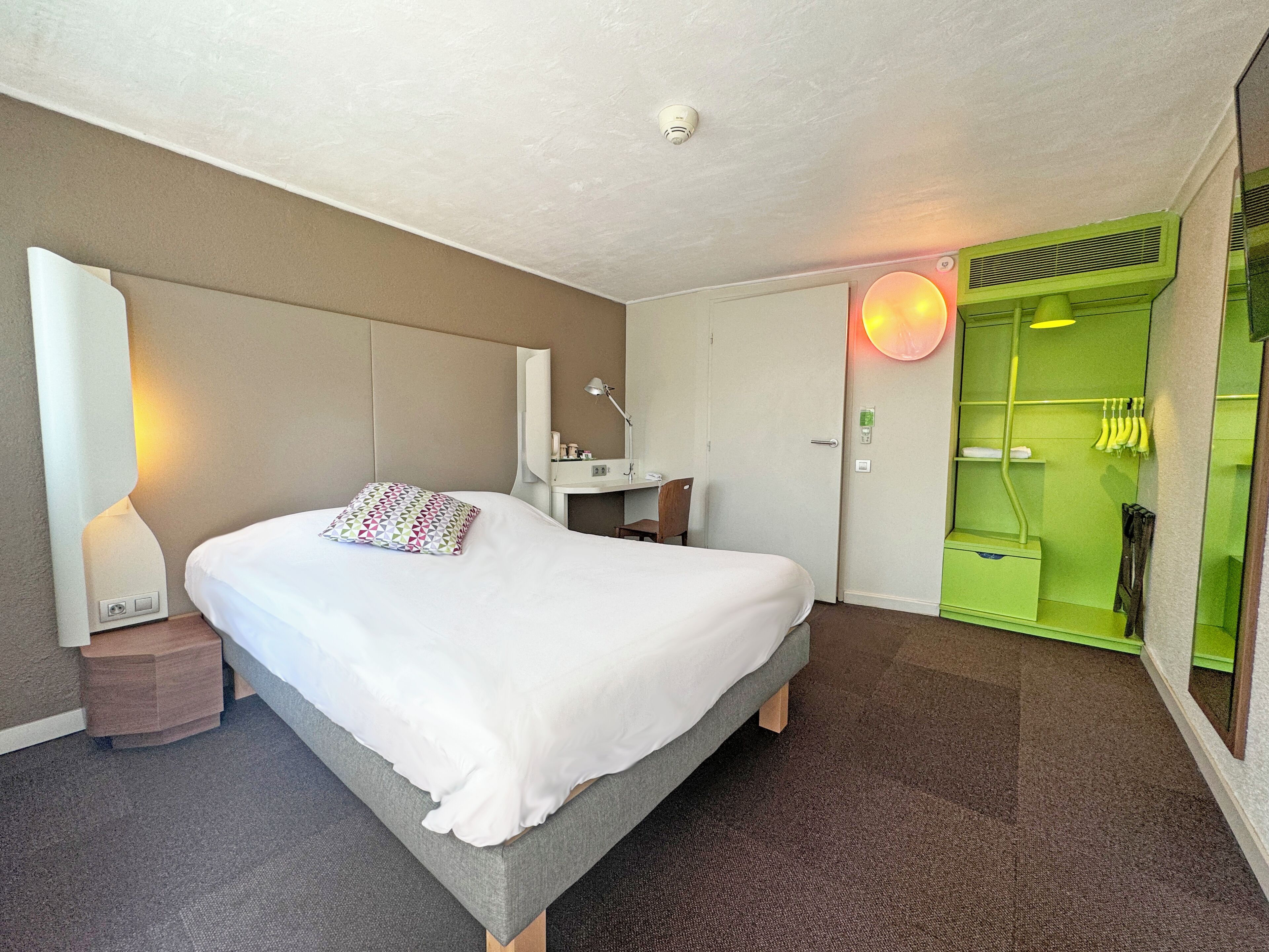 Una habitación de hotel con cama, escritorio, espejo, armario verde y lámpara fijada a la pared.