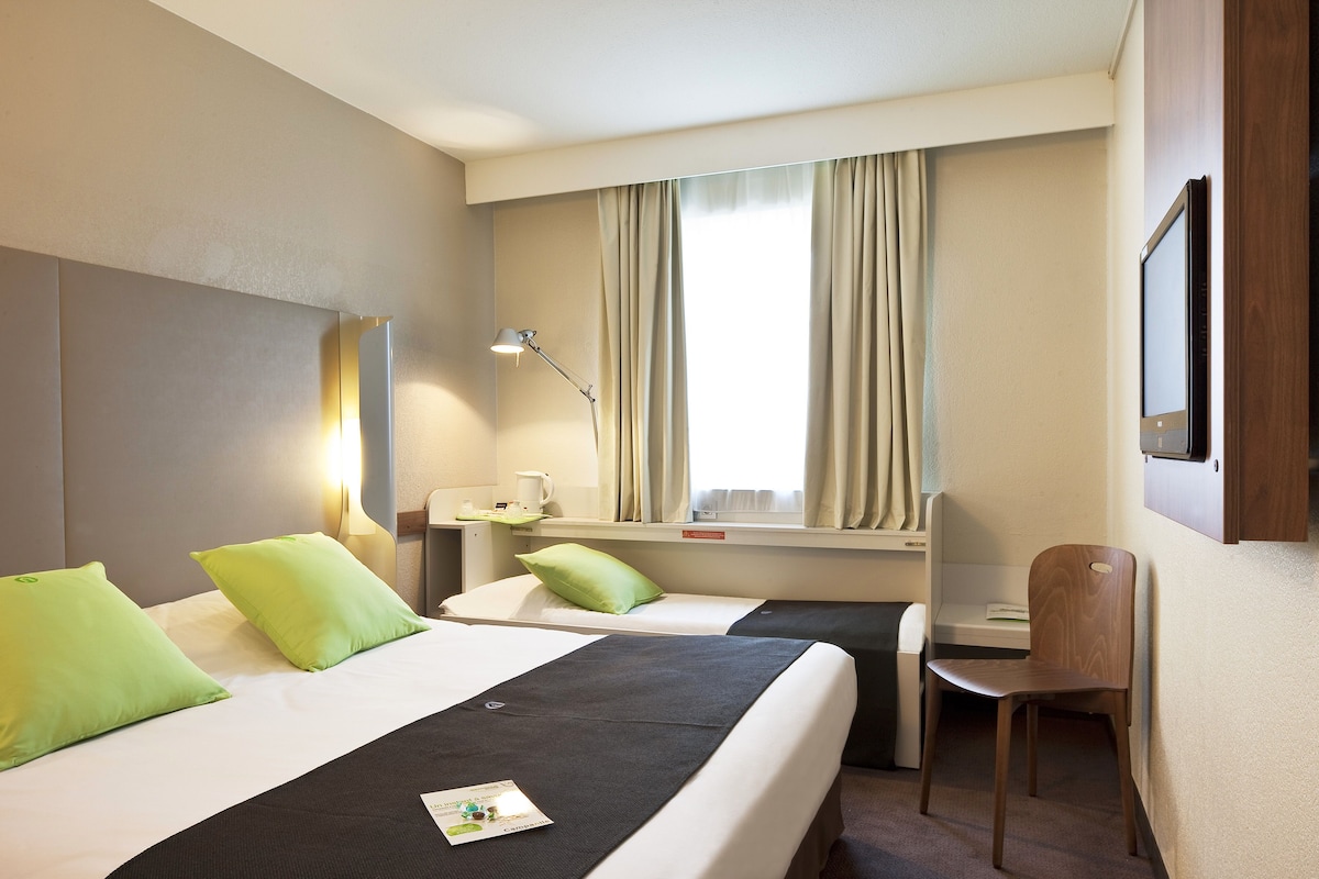 Habitación de hotel con dos camas, un escritorio, una silla, una lámpara y una ventana con cortinas.