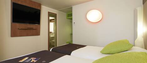Habitación de hotel con dos camas, un televisor montado en la pared, una mesita de noche con una revista y una lámpara de pared redonda.