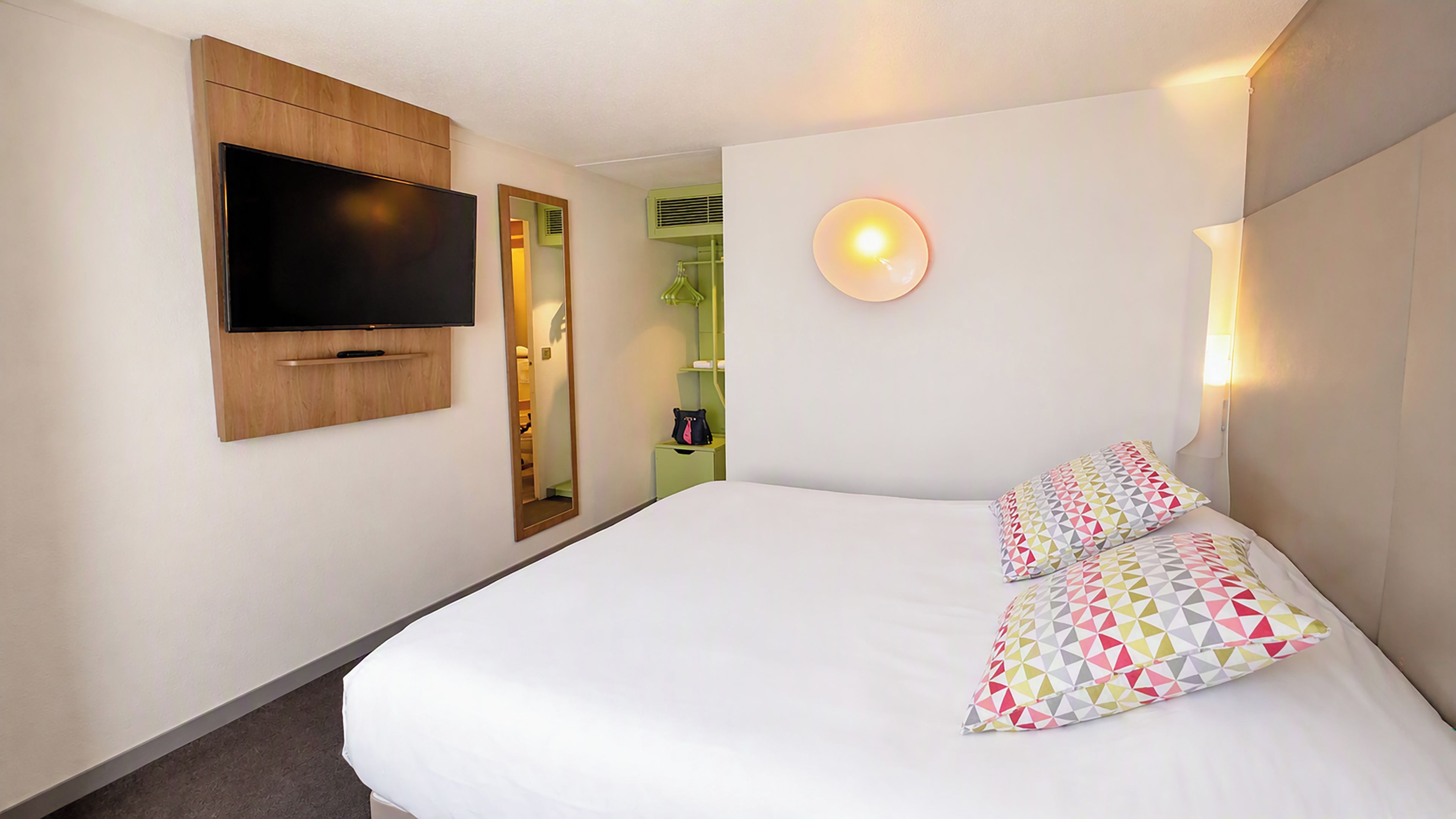 Una habitación de hotel con una cama, un televisor de pantalla plana montado en la pared, una mesita de noche y una lámpara de pared.