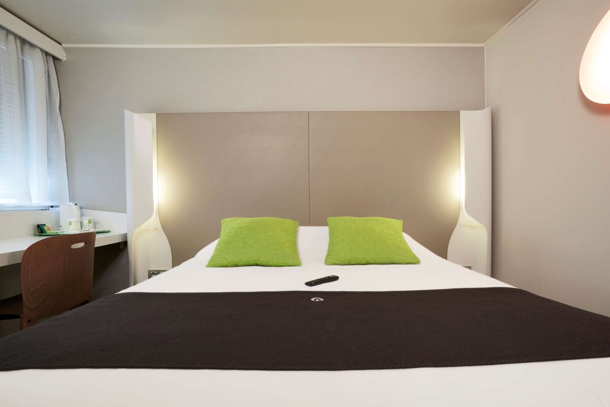 Een hotelkamer met een bed, twee groene kussens, een afstandsbediening, een bureau en een stoel.